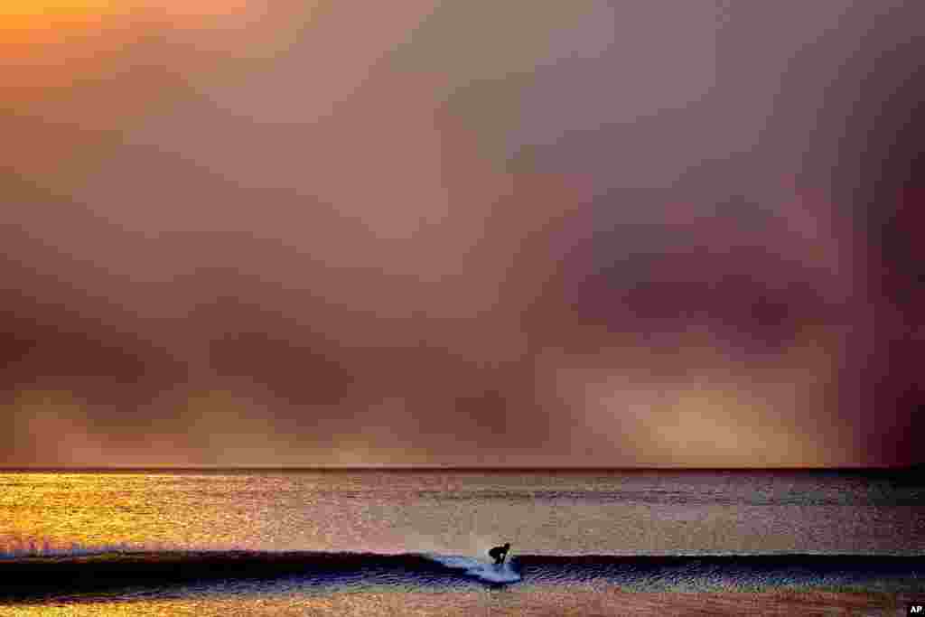 Um surfista levanta em Santa Monica, Califórnia, durante o pôr do sol sob um céu enegrecido pelo incêndio de Palisades em Pacific Palisades, 7 de janeiro de 2025.